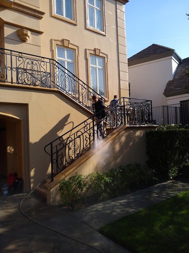 A person power washing the stone steps of a tan house with ornate black metal railings.