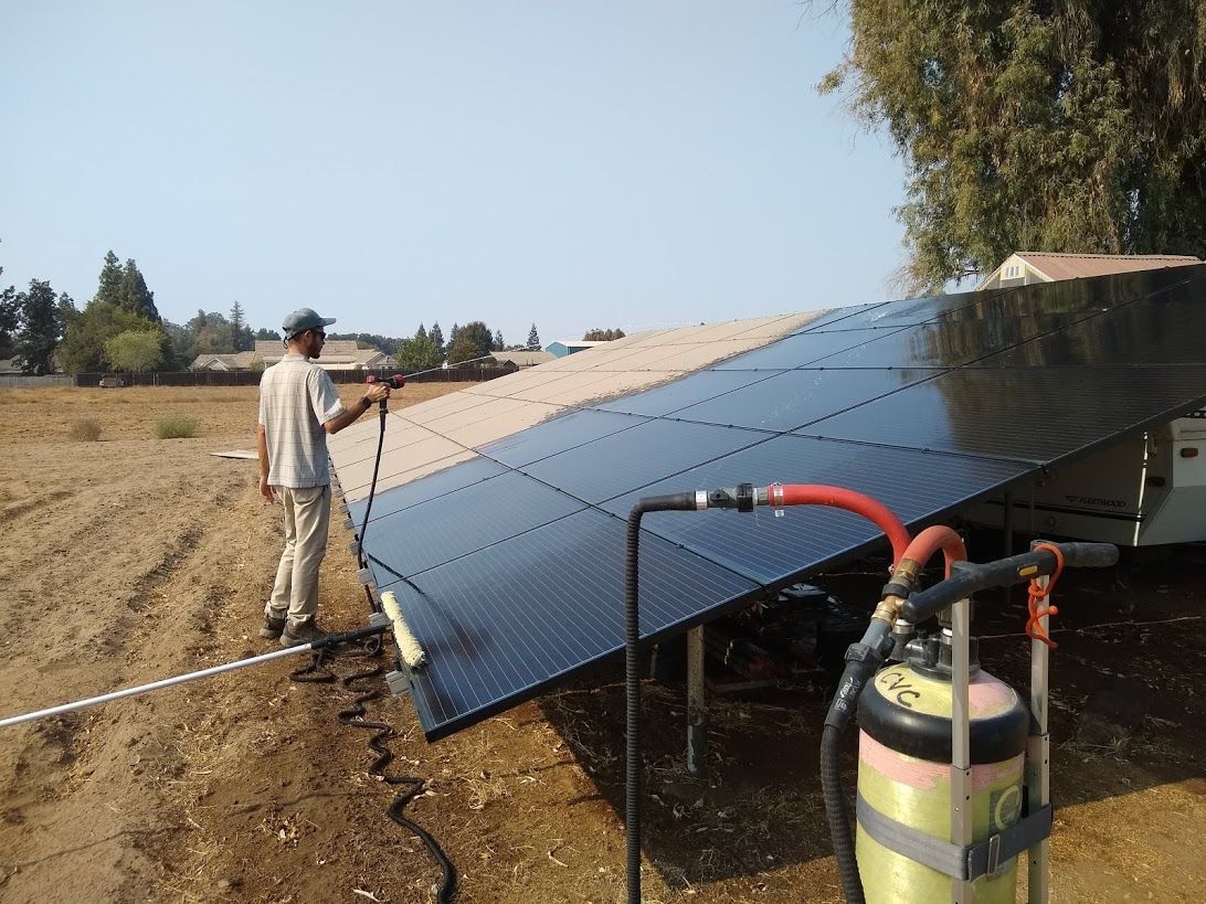 A person cleans ground-mounted solar panels in a field using a hose and sprayer attached to a portable tank.
