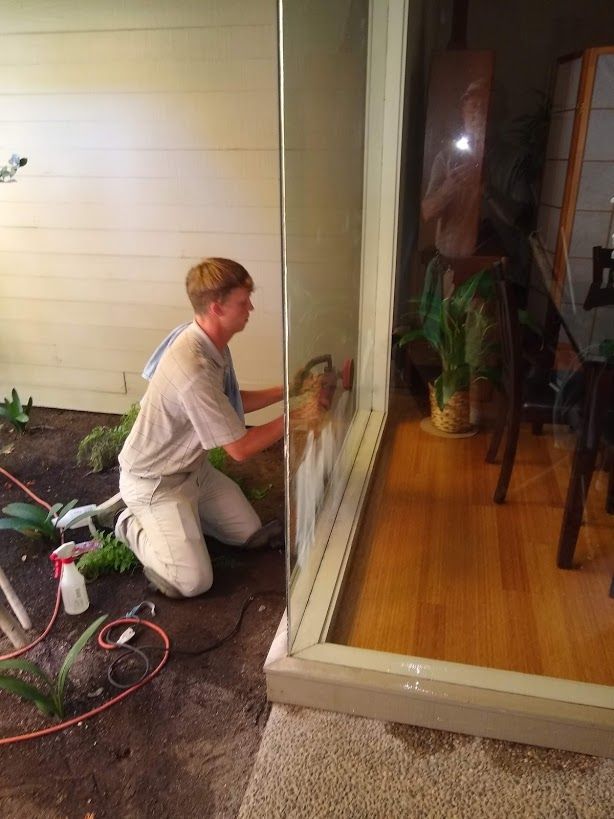 A person kneeling on soil cleans a large glass window that separates an outdoor garden area from an indoor wood-floored room.