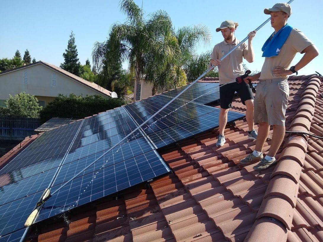 Two people cleaning solar panels on a tiled residential roof using a long-handled brush.