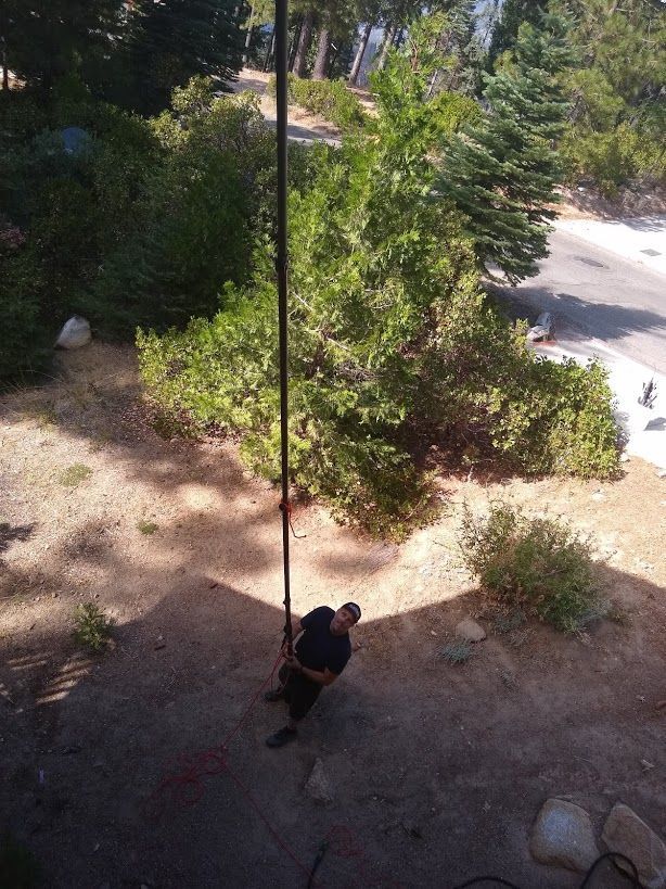 A person in dark clothing stands on a dirt ground holding a long, vertical pole reaching up toward the camera.