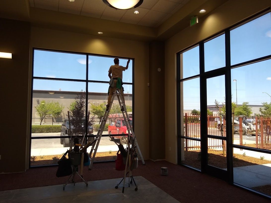 A person stands on a tall, A-frame ladder, cleaning the large floor-to-ceiling windows of a sunlit building interior.