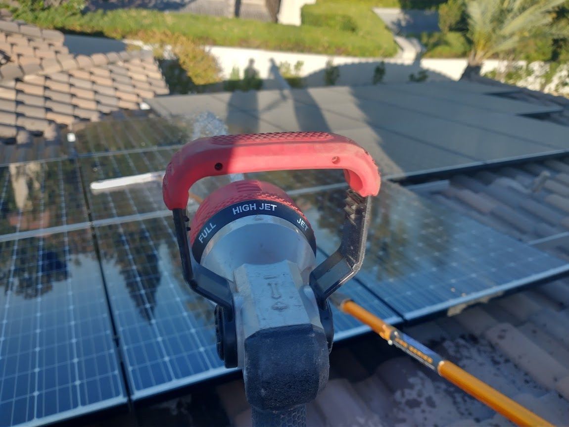 A red garden sprayer head held over rooftop solar panels, with a pole extending to the right.