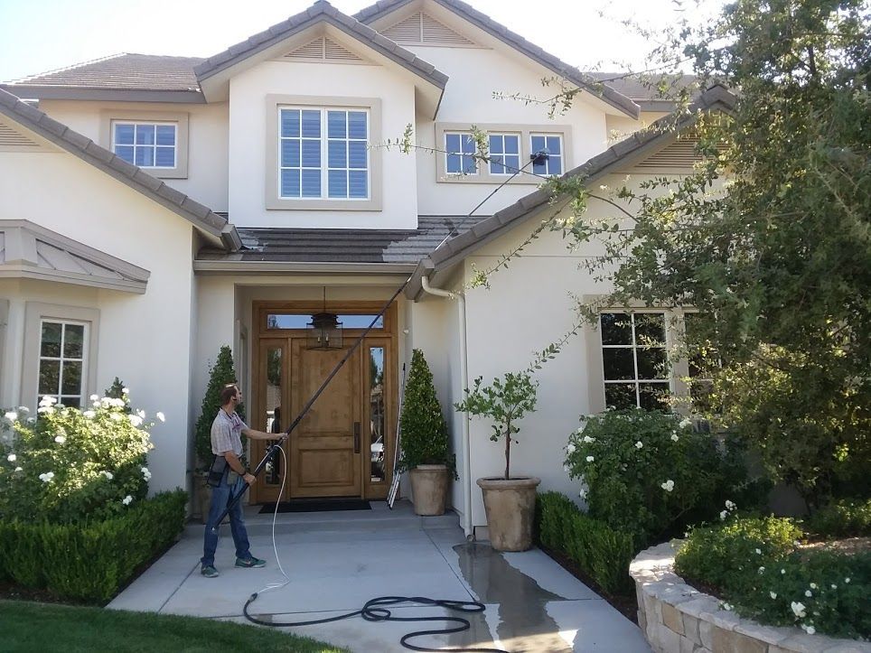 A person uses a long water-fed pole to clean the exterior windows of a two-story beige house with a wooden front door.