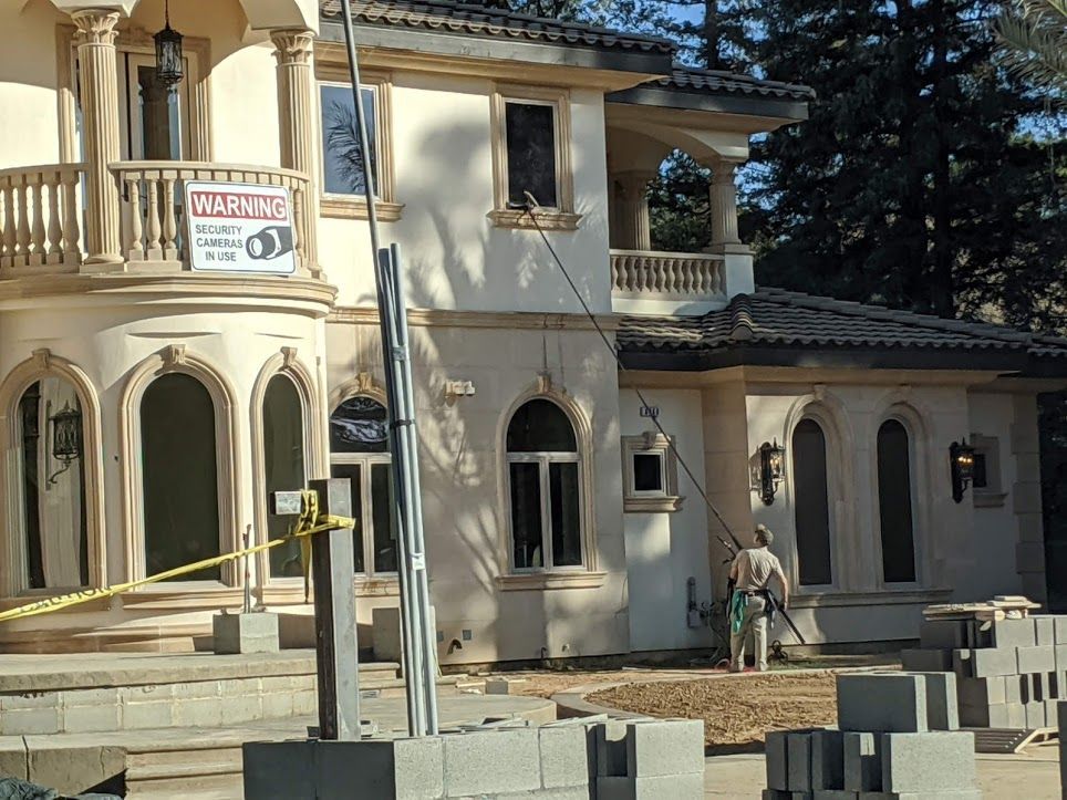 A worker in a beige uniform uses a long pole to clean the exterior second-story window of a large, light-colored house.