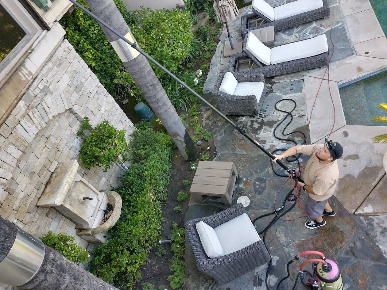 A person standing on a stone patio uses a long extension pole to clean the exterior of a multi-story stone home.