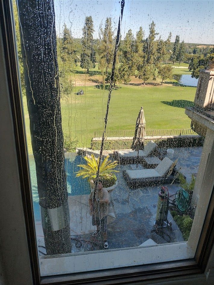 A person on a patio cleans a large window, reflecting the pool area and a green golf course in the background.