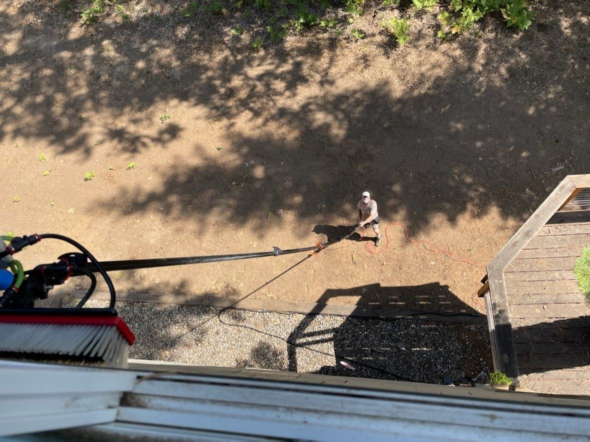 A person stands on the ground below, extending a long pole with a cleaning brush attached to reach an upper-story window.