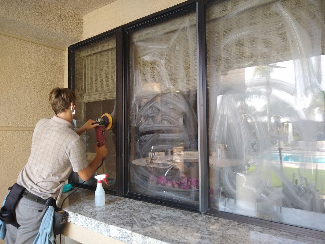 A person uses a power polisher on a window covered in white residue, standing at a stone countertop on a patio.