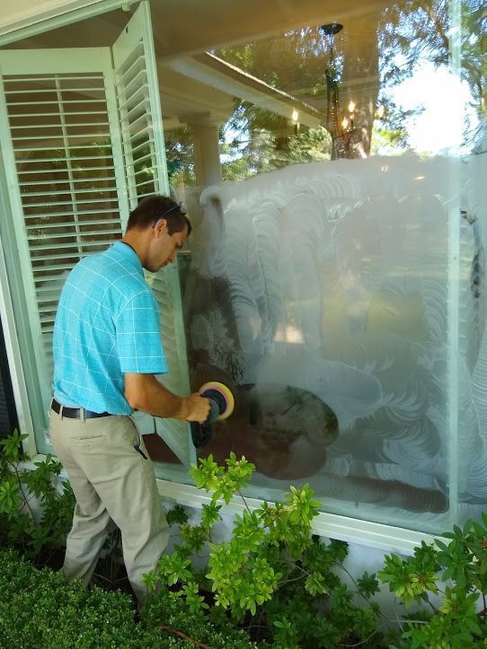 A person in a blue polo shirt uses a rotary tool to polish or buff a cloudy patch on a large residential window.