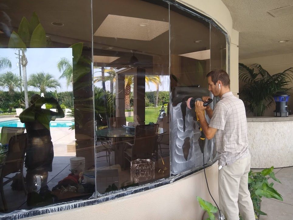 A person uses a power tool to apply tinting film to a curved, outdoor glass wall overlooking a swimming pool.
