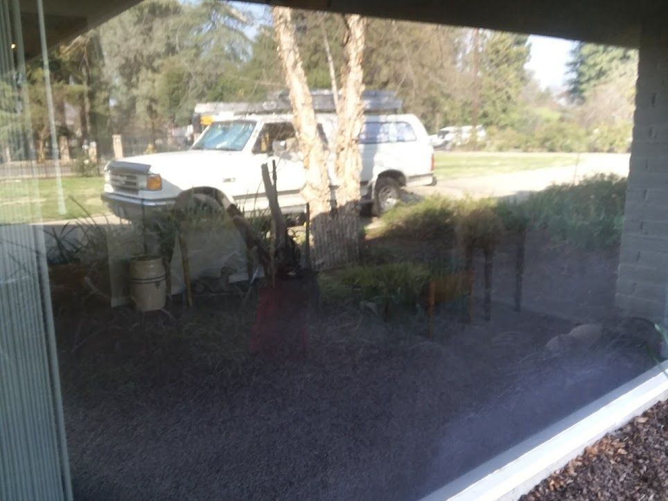 A view through a window showing a white truck parked on a gravel driveway in front of trees and a green lawn.