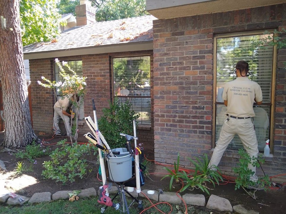 Two workers in tan uniforms clean windows on the exterior of a brick house with cleaning tools and equipment nearby.