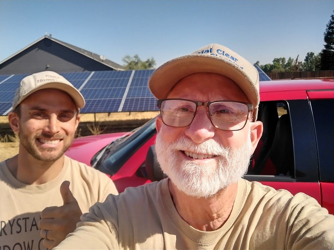 Two people in tan shirts and caps pose for a selfie in front of a red vehicle and a ground-mounted solar array.
