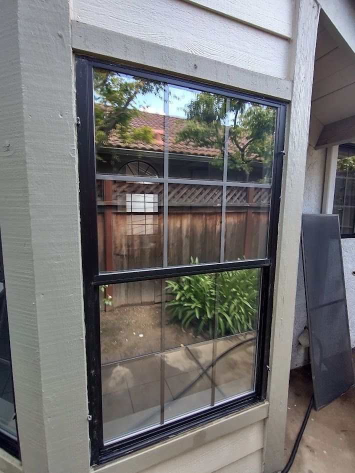 A black-framed single-hung window with a grid pattern on the exterior of a building with beige siding.