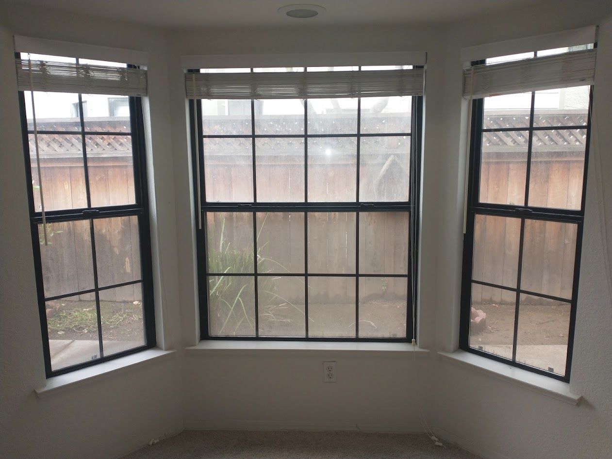 A three-paneled bay window with black frames and slatted blinds, overlooking a wooden fence and yard.