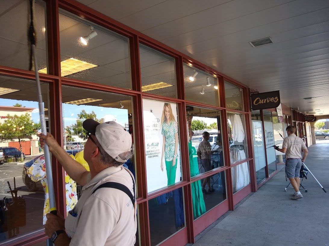 Two window cleaners use long-handled poles to clean the exterior glass of storefronts under a covered walkway.