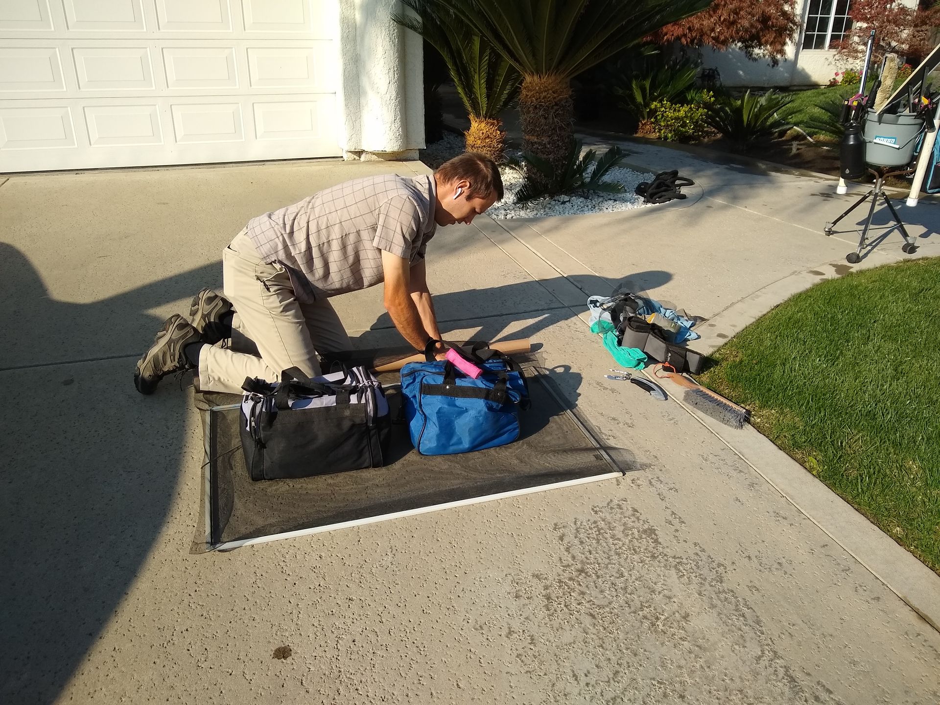 A person kneels on a driveway next to a blue duffel bag, a black bag, and some scattered equipment in front of a garage.