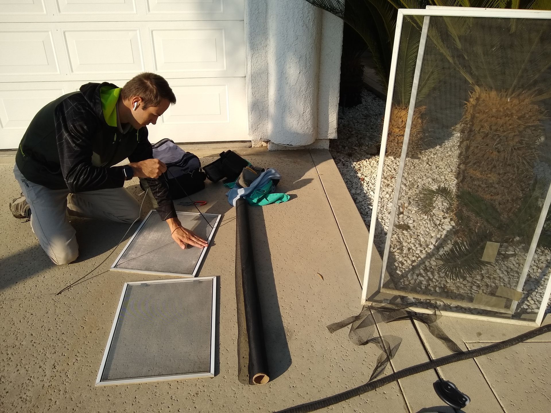 A person kneeling on a concrete driveway, working on replacing screen mesh into white window screen frames.