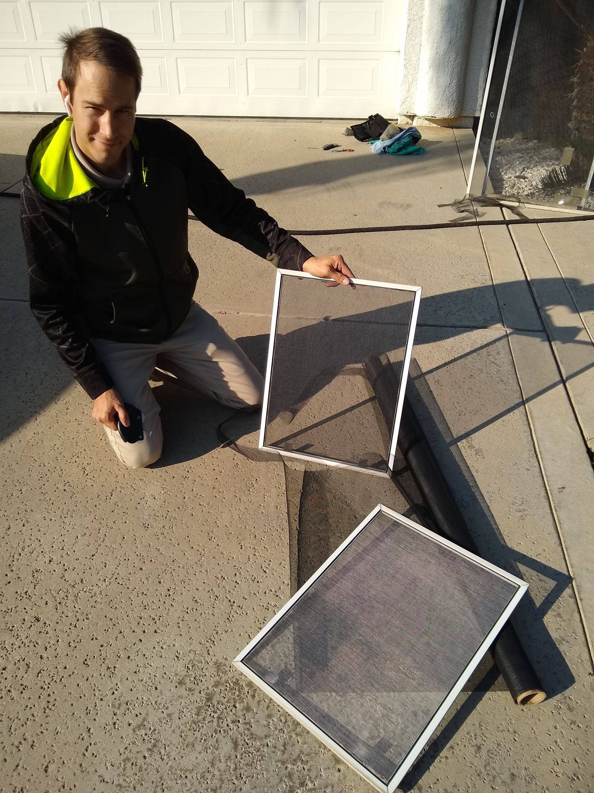 A person kneels on a driveway, holding a window screen frame near another completed screen and a roll of mesh.
