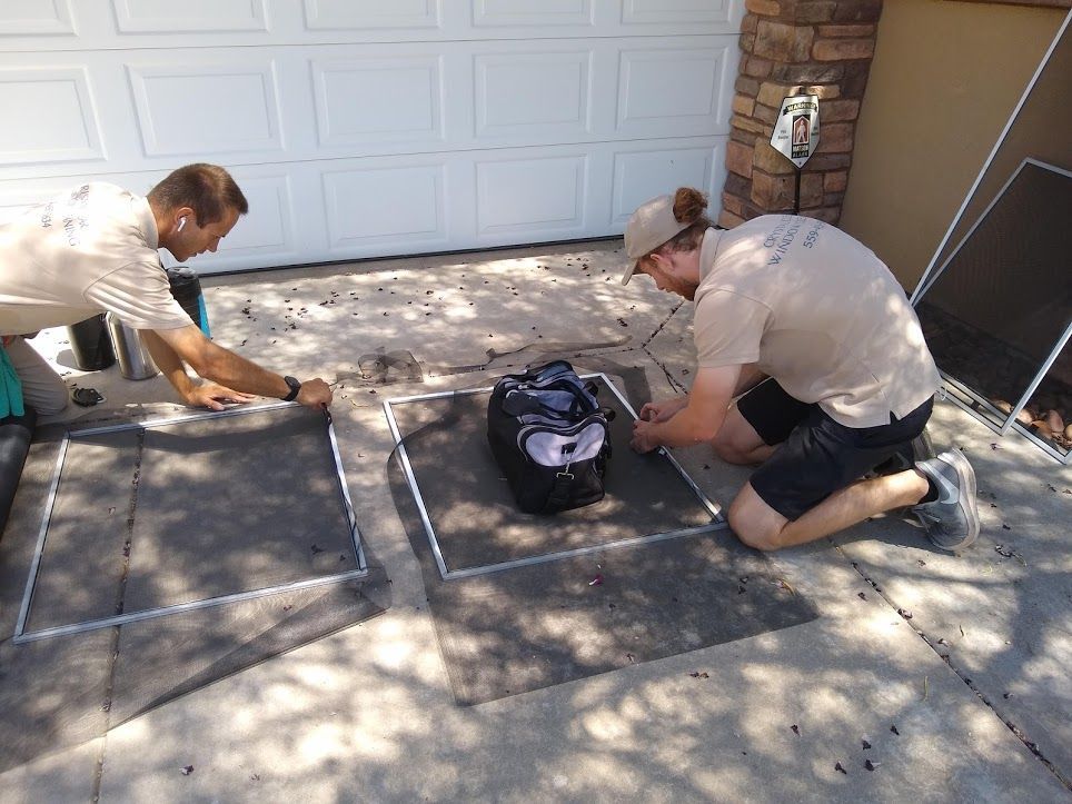 Two technicians in tan uniforms assemble window screens on a driveway in front of a closed garage.