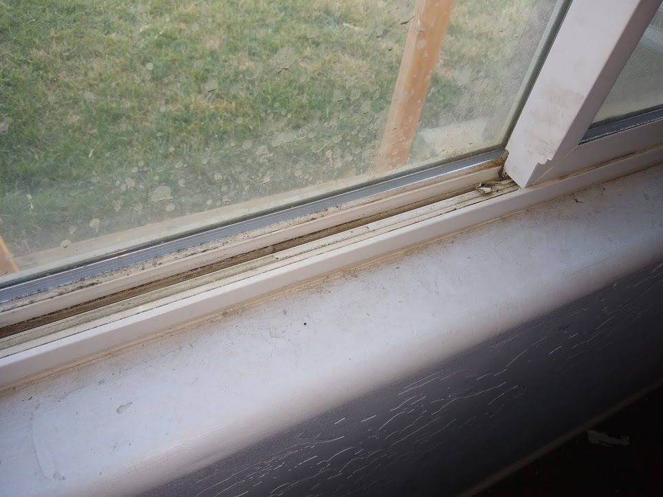 A close-up view of a dusty window sill and the bottom track of a sliding vinyl window frame.