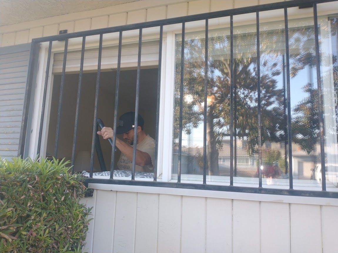 A person wearing a dark cap inside a house, visible through a window with black security bars.