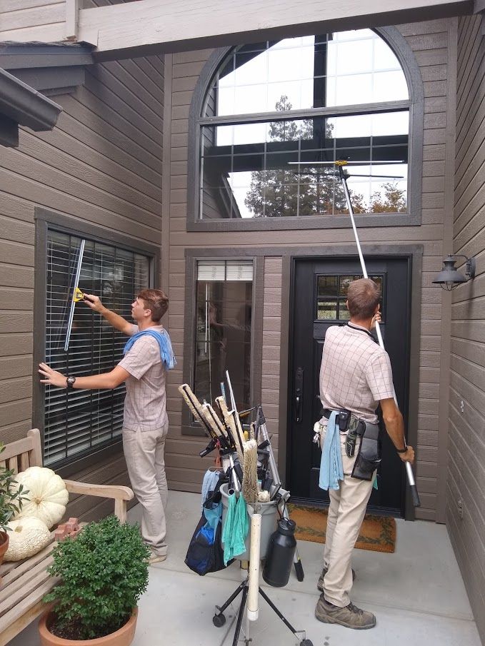 Two workers cleaning the windows of a brown home exterior using long-handled squeegees.