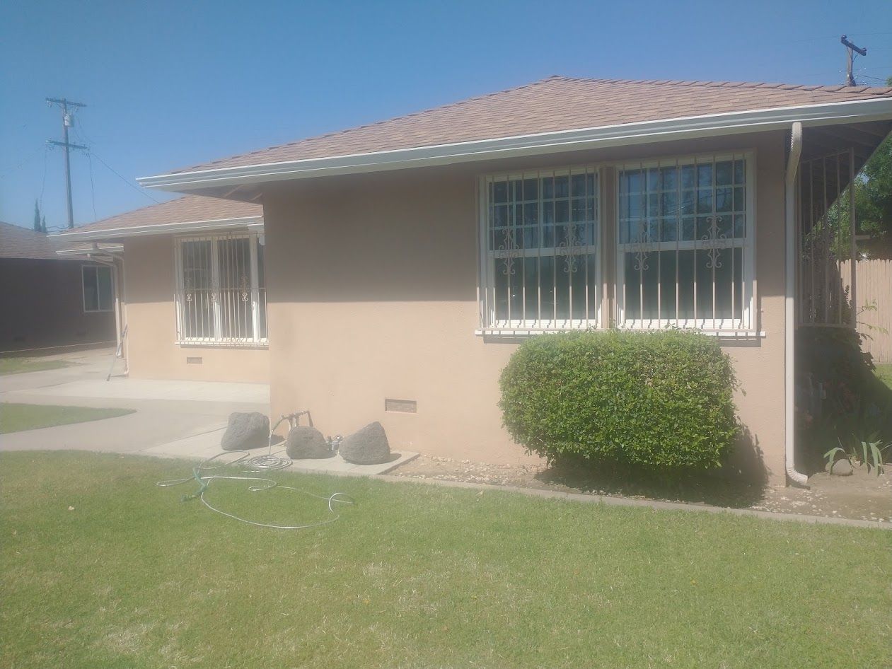 A single-story, tan-colored house with a brown shingled roof, windows with security bars, and a trimmed green bush.
