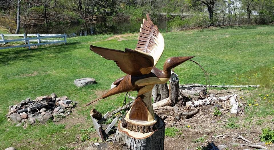 Brown bird sculpture on a stump in a grassy yard, wings spread near a fire pit and stone border.
