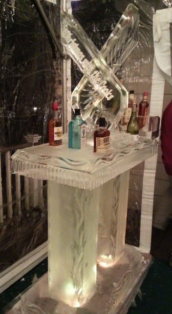 Ice sculpture centerpiece on a lit table with several bottles and drinks at an event.