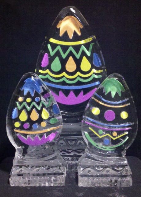 Three decorated glass Easter eggs with colorful zigzag and dot patterns on clear stands.