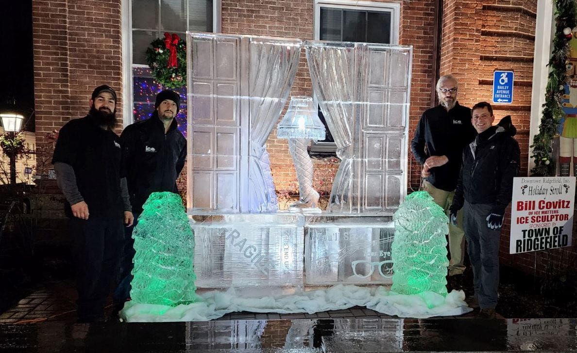 Person posing in a clear ice sculpture frame at night, flanked by two people and green-lit ice pillars