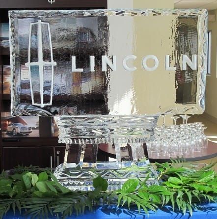 Clear ice sculpture with Lincoln logo on a decorated table, surrounded by green leaves