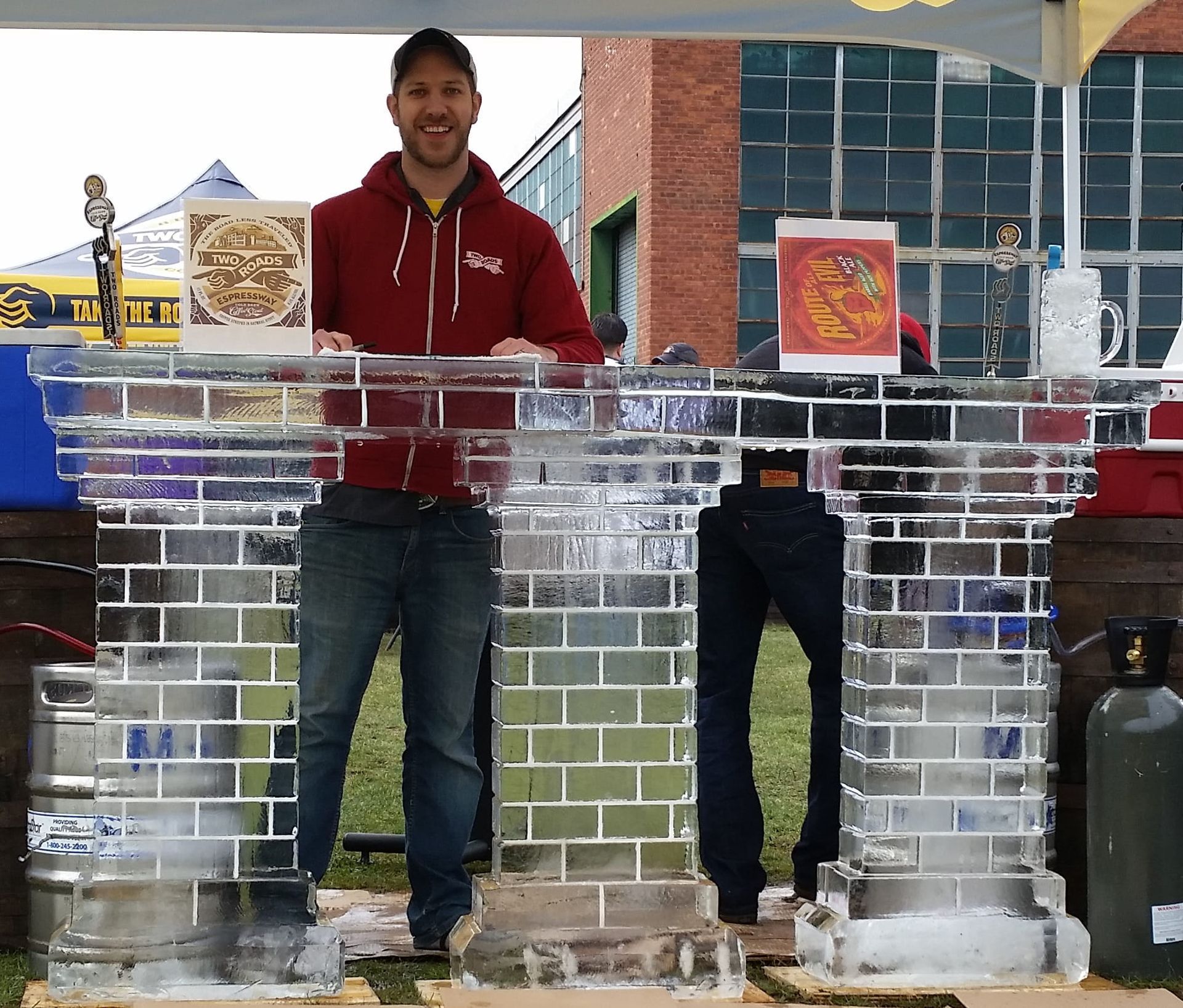 Two people behind an ice bar at an outdoor event, one in a red hoodie, with drinks and equipment nearby.