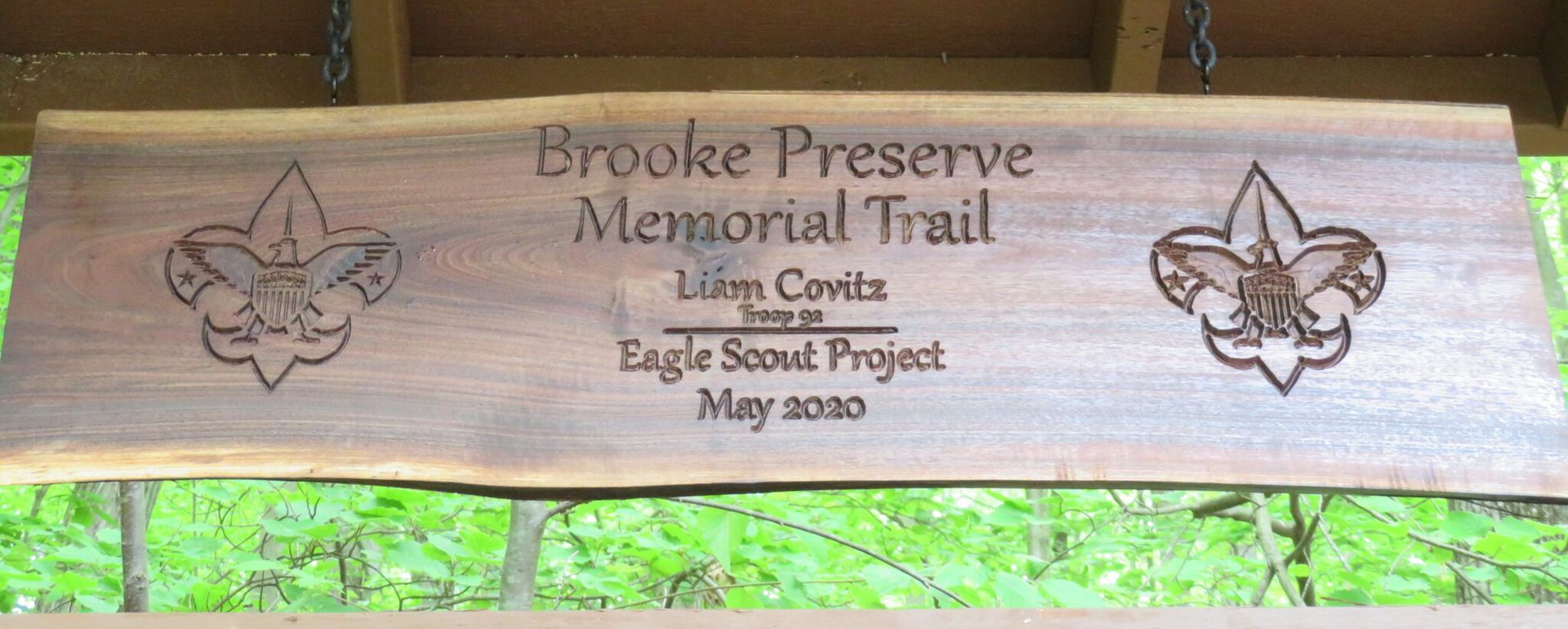 Wooden sign reading “Brooke Preserve Memorial Trail” with Eagle Scout Project and May 2020, framed by trees.