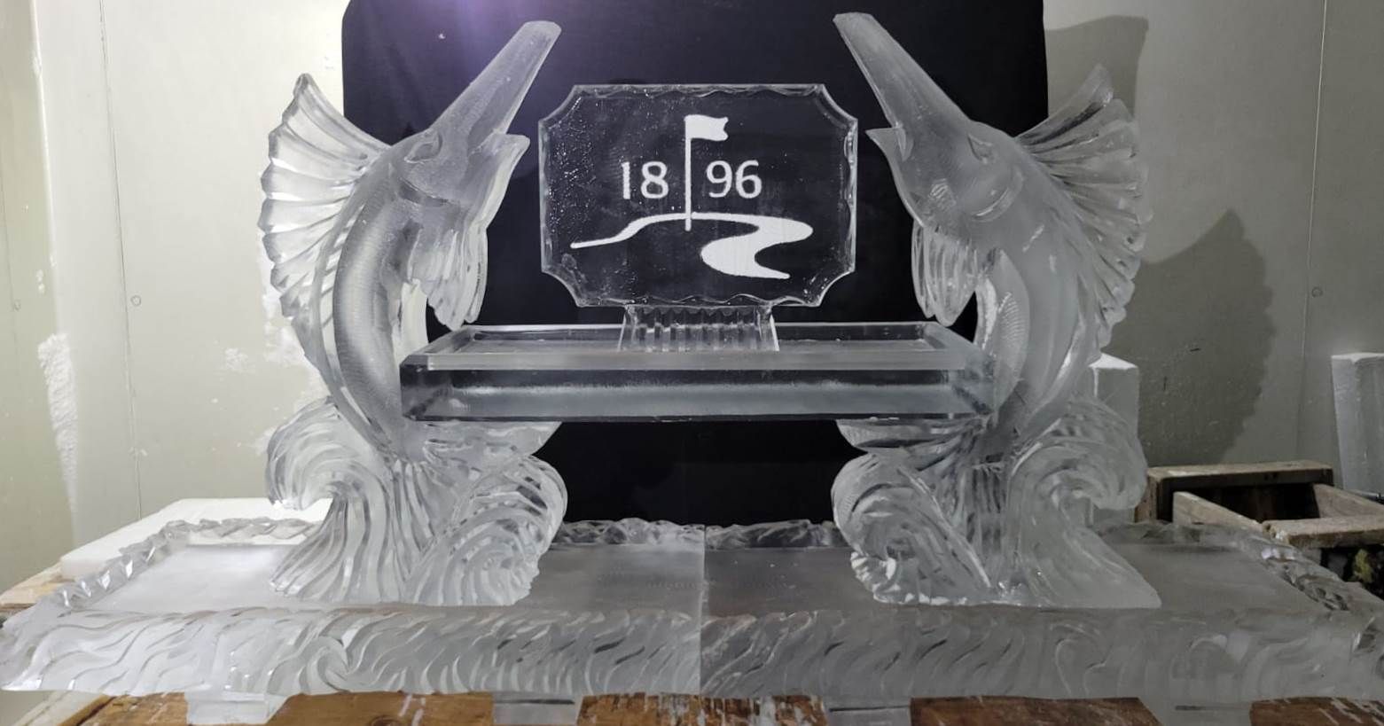 Clear ice sculpture with wings framing an “1896” book emblem on a wooden table