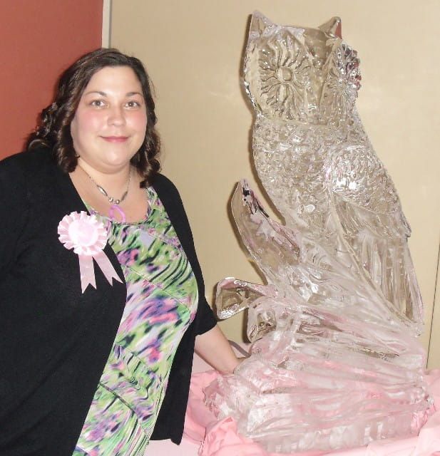 Woman standing beside a clear ice sculpture indoors, wearing a black cardigan and floral dress with a pink ribbon.