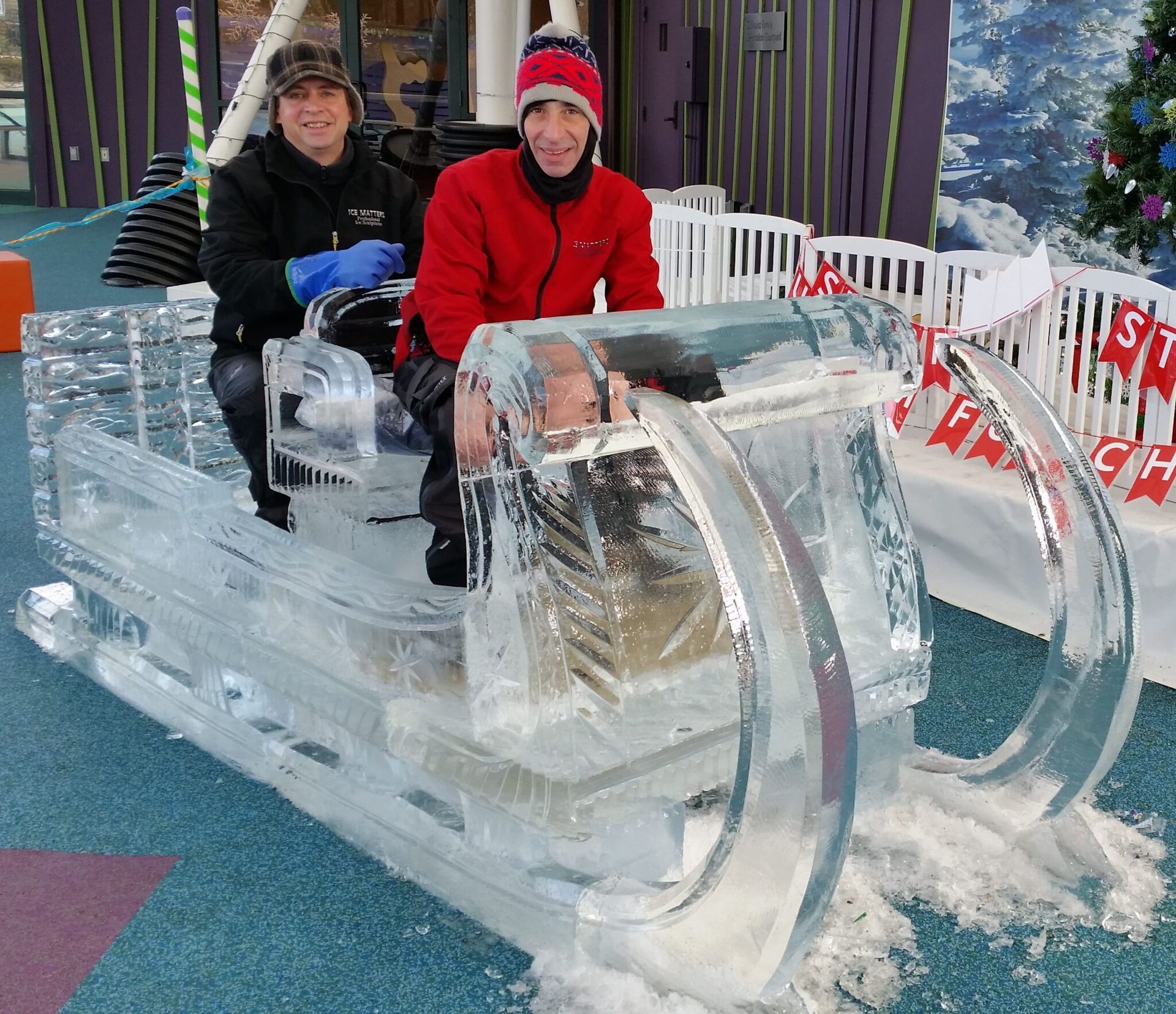 Two people sit in an ice sculpture sled at a snowy outdoor event