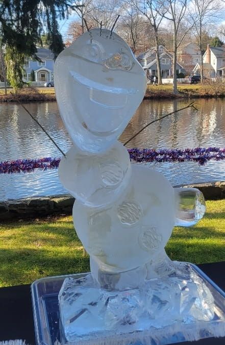Clear ice sculpture on a stand beside a canal, with houses and trees in the background.