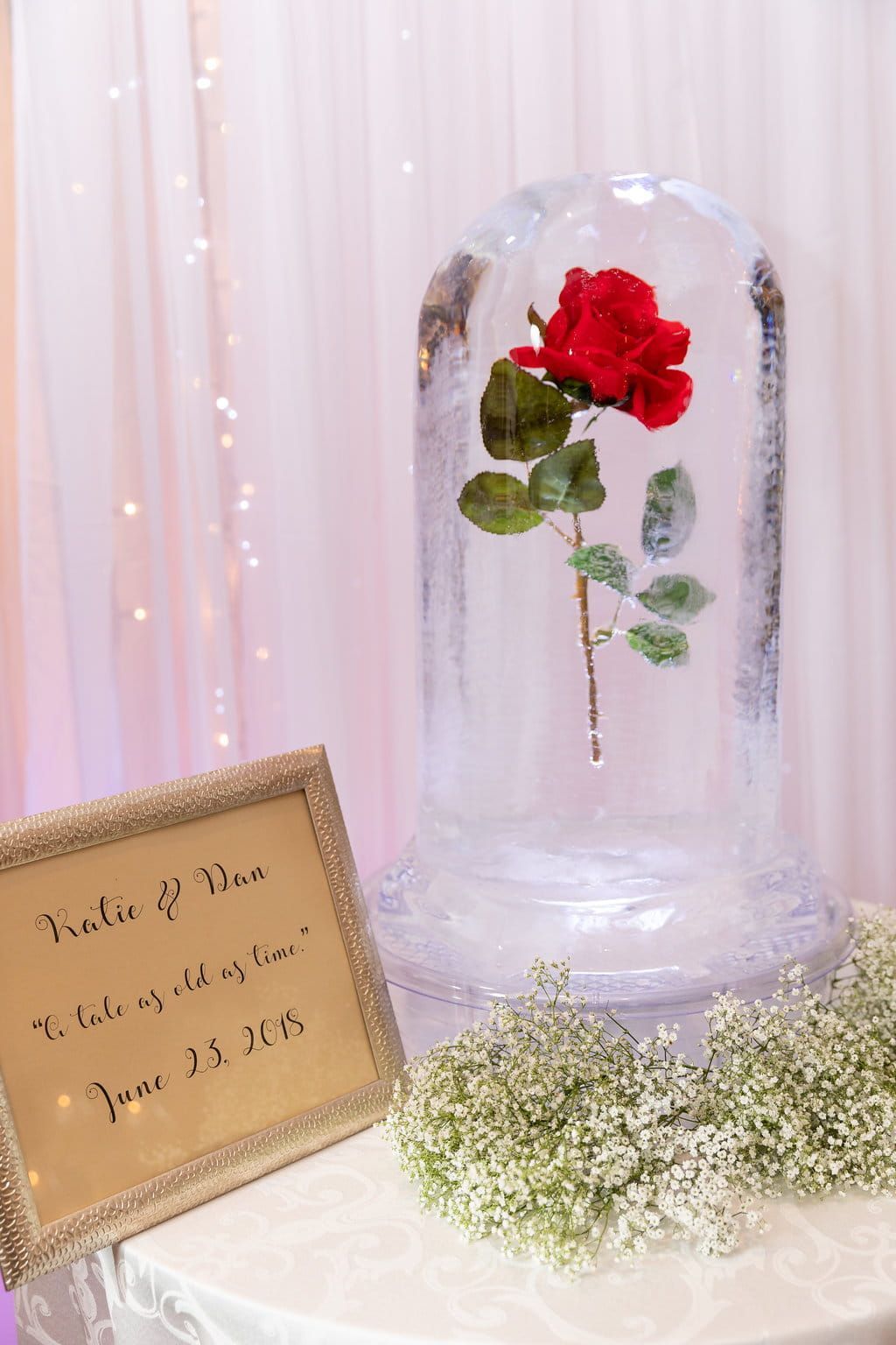 Single red rose in a clear glass dome beside a small plaque and white flowers on a pink backdrop