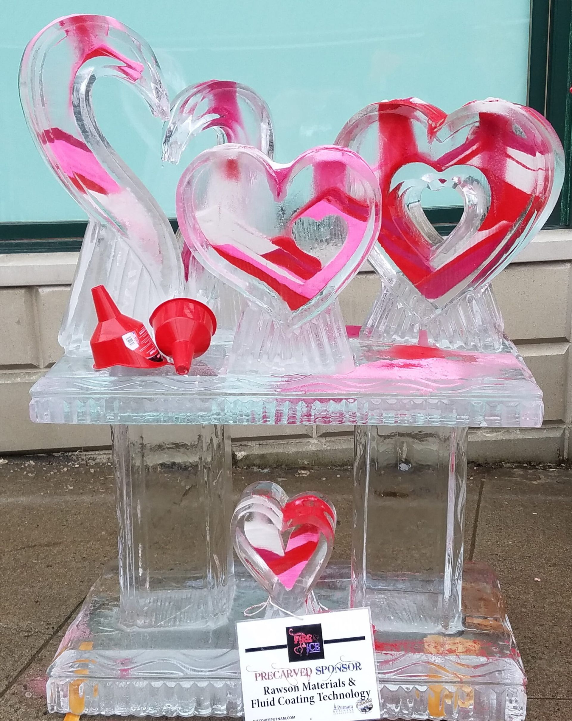 Pink heart-shaped ice sculpture on a clear stand with red accents, displayed indoors near a wall.