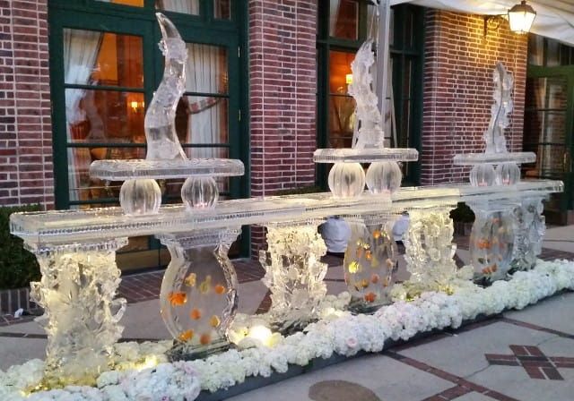 Clear ice sculptures and fountain display on a brick patio with white floral decorations and warm lights