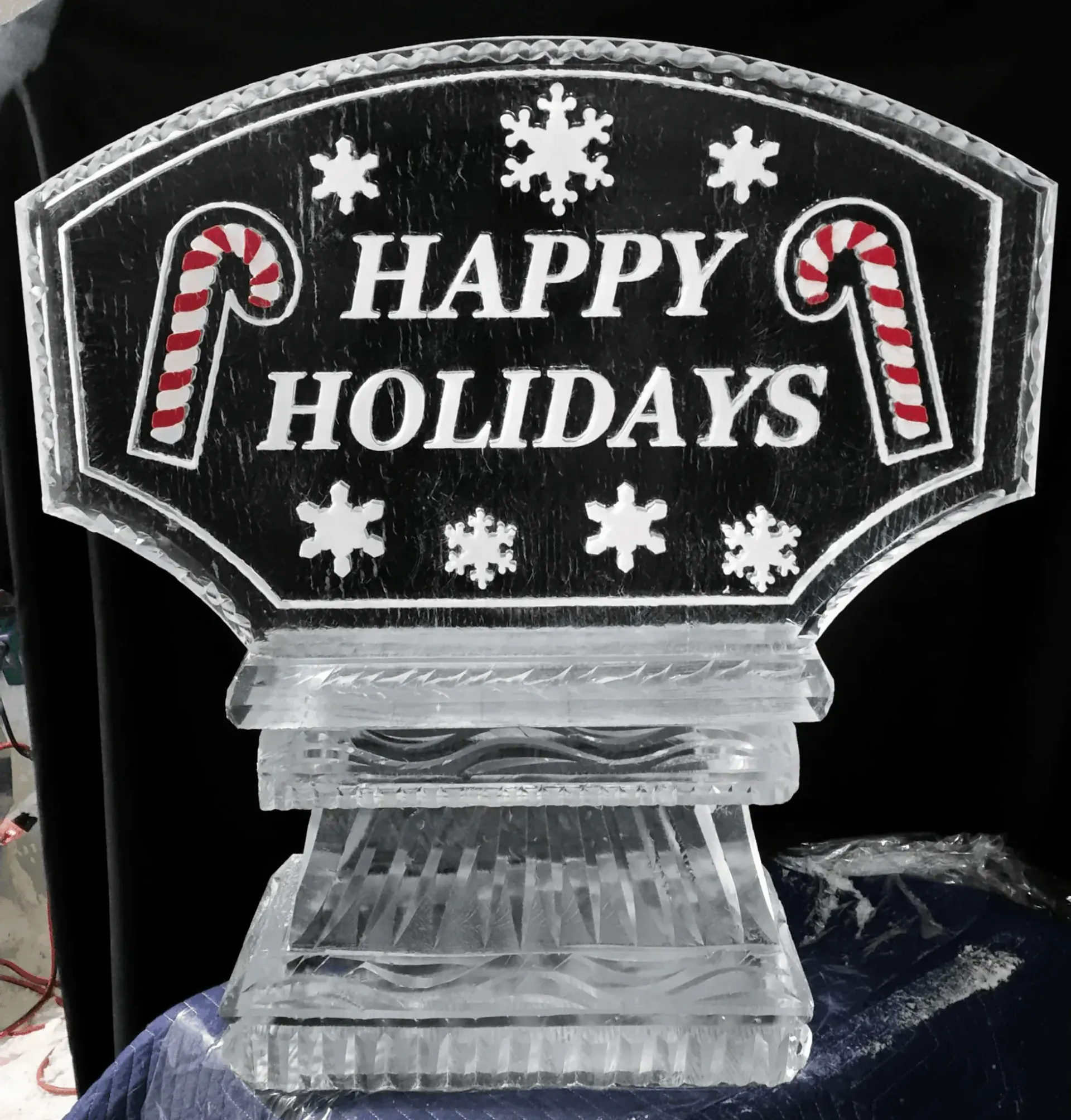 Clear ice holiday sign reading “Happy Holidays” with candy canes and snowflakes on a black background