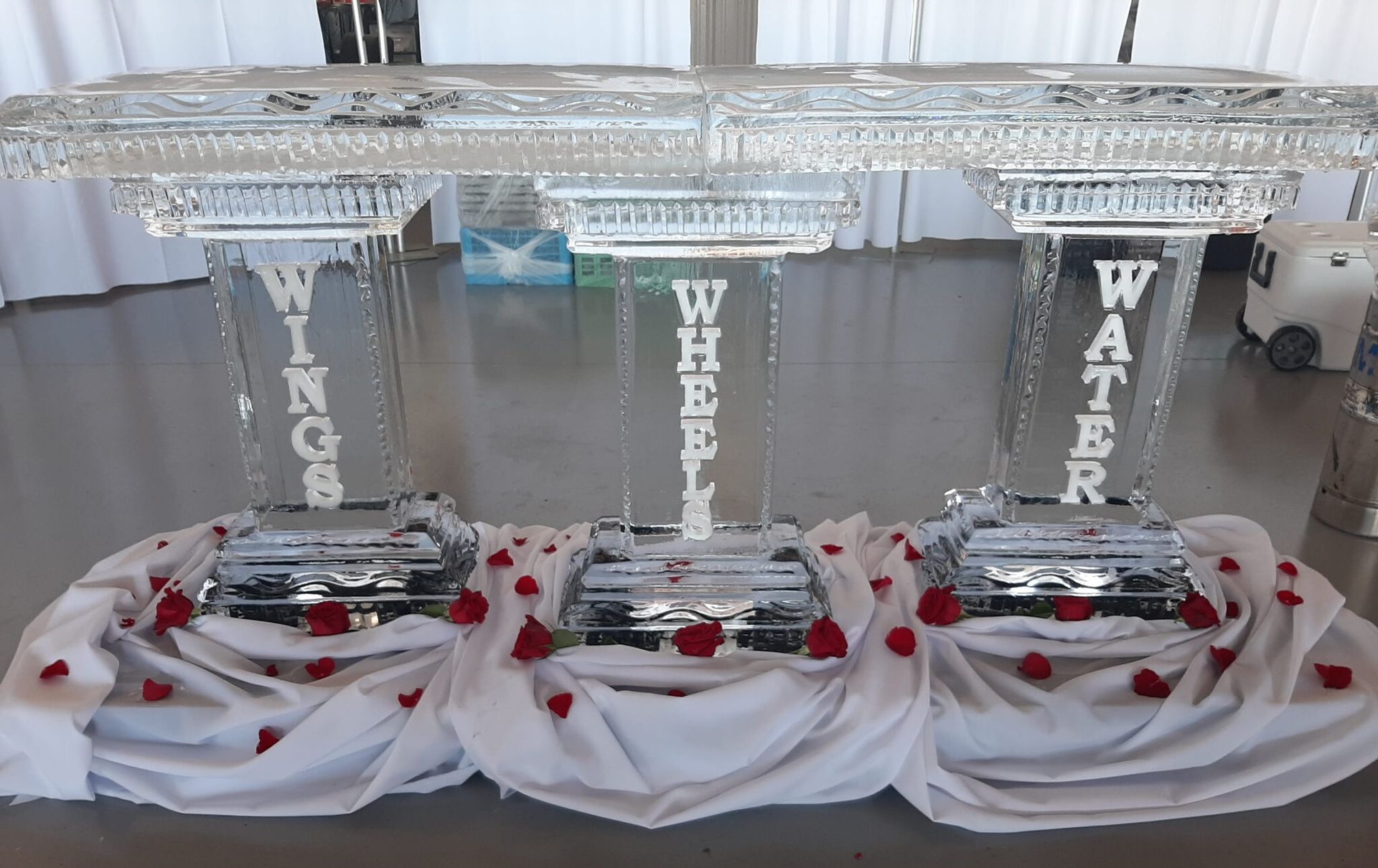 Three clear acrylic memorial podiums on white draped tables with red roses and silver plates.