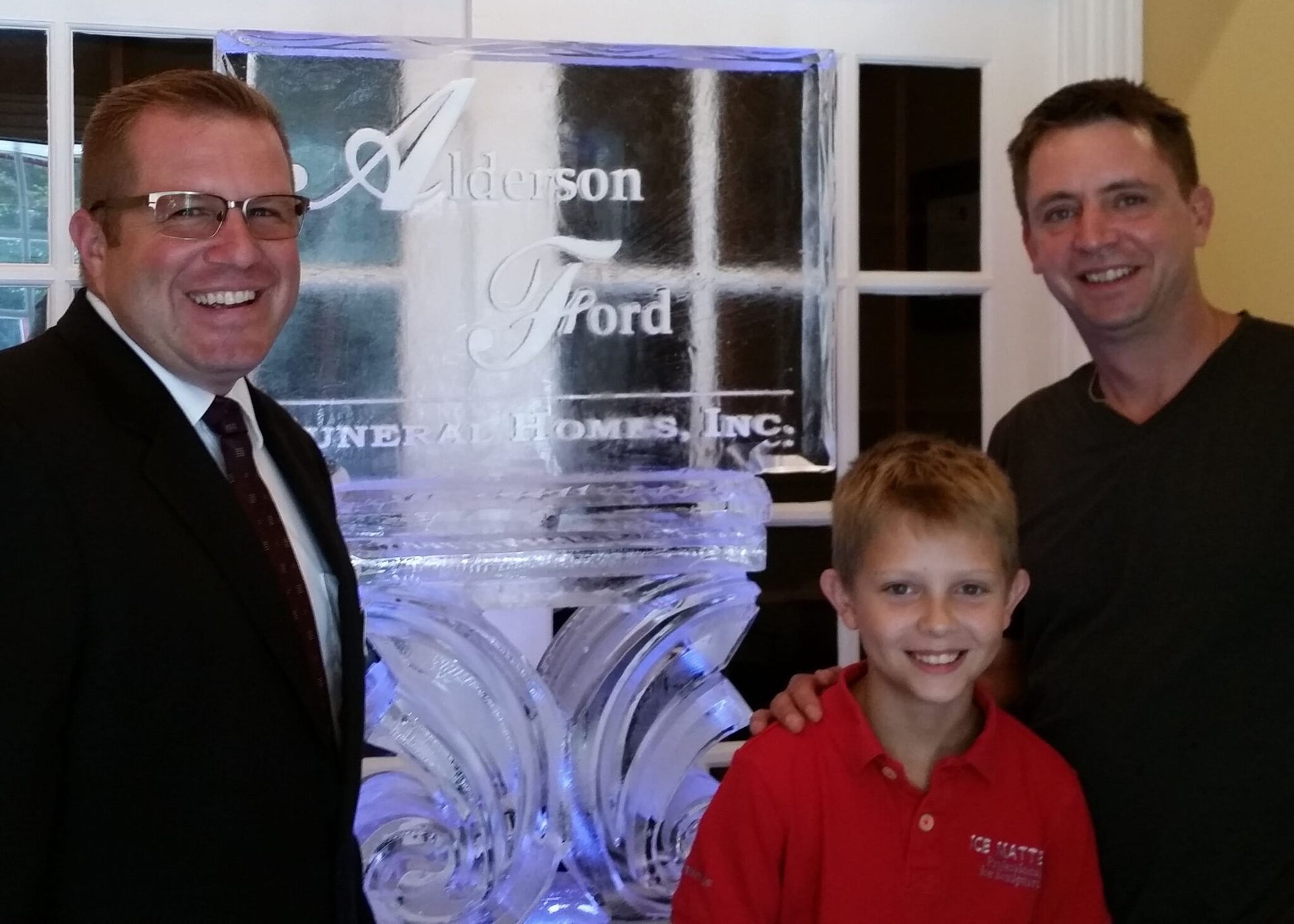 Three smiling people standing indoors beside an etched glass sign and trophy.
