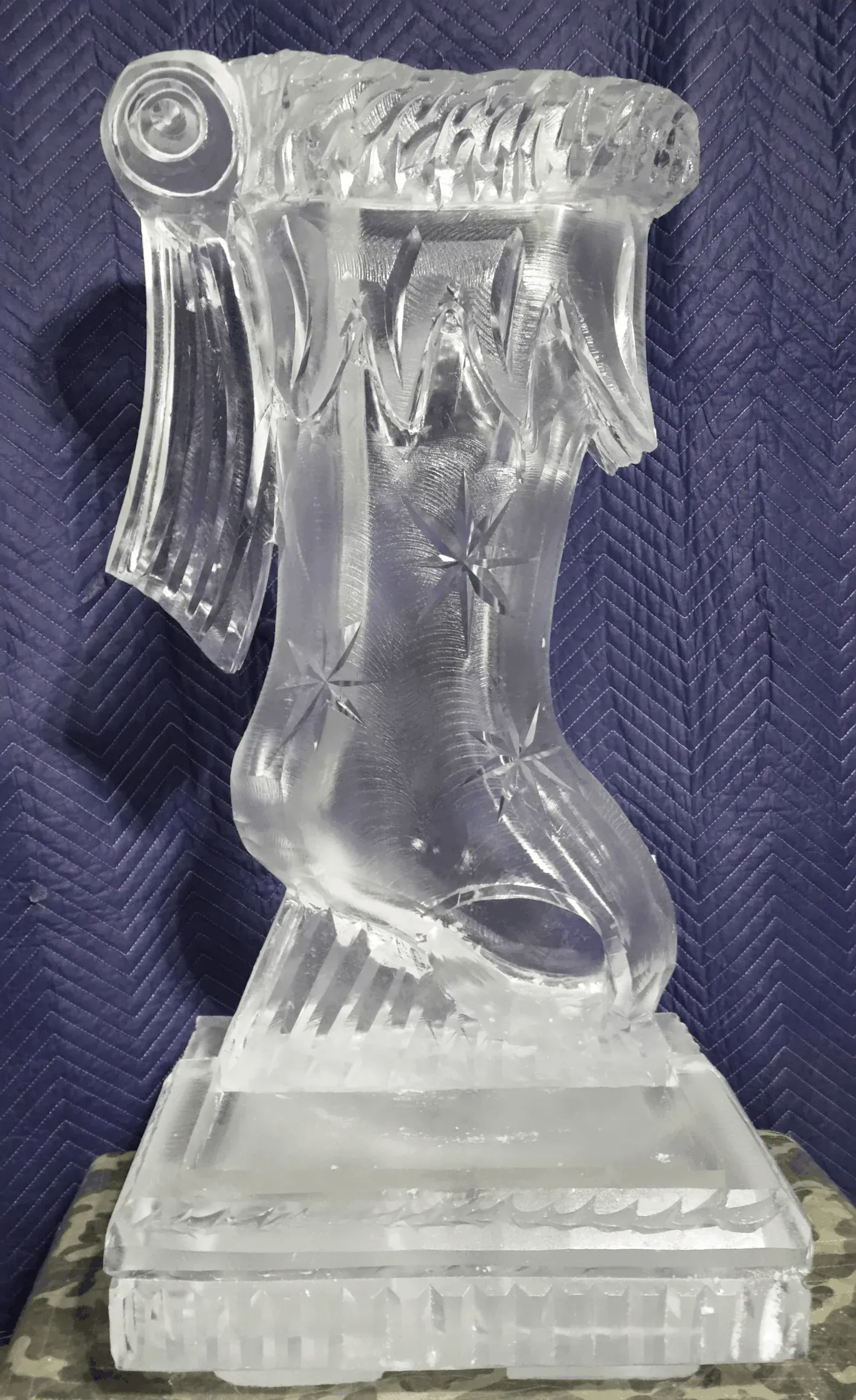 Clear glass figurine of a winged female figure on a pedestal, viewed from the front.