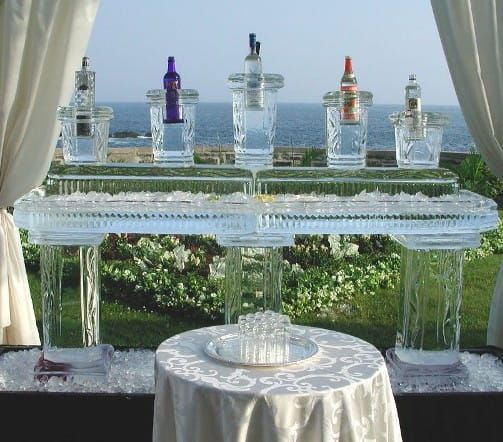 Decorative glass beverage dispensers and cups on a clear glass table at an outdoor event