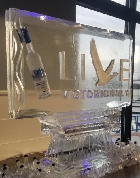 Clear ice sculpture with “LIVE VICTORIOUSLY” text and a hanging bottle, lit with purple-blue accents.