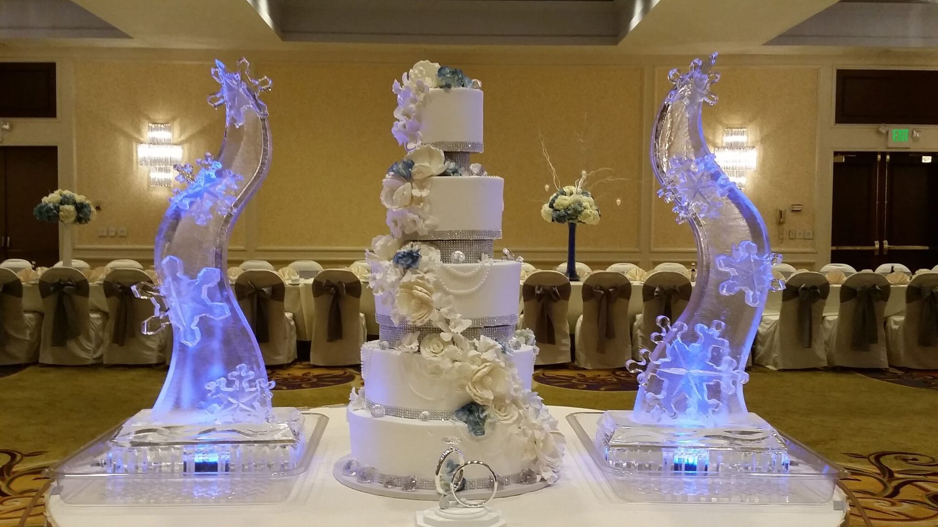 Elegant white tiered cake with flowers, flanked by two glowing blue decorative swans on a banquet table.